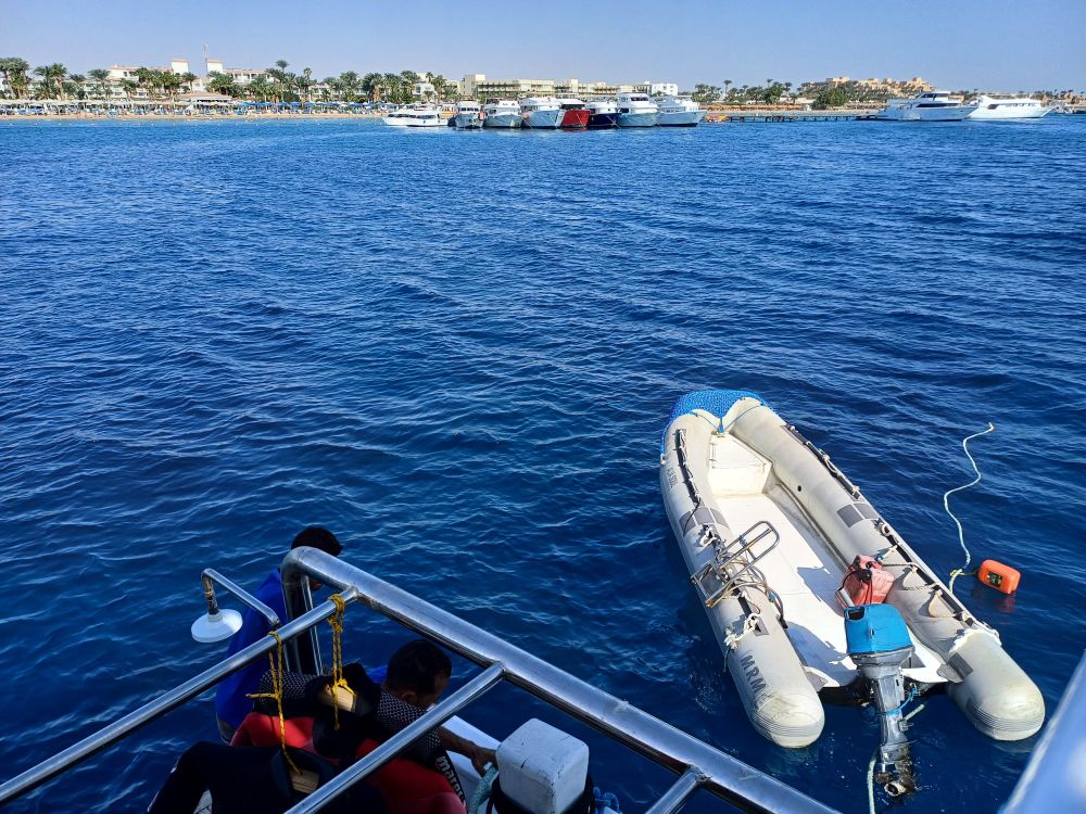 Blick von einem Boot auf dem Meer.
Ein Schlauchboot ist daneben zu sehen.
Vom Boot zum Propeller des Schlauchbootes und noch etwas weiter  ist eine dicke Leine, mit orangenem Schwimmkörper zu sehen.
Im Hintergrund: viele andere Boote, eng nebeneinander, jeweils mit dem Bug zum Betrachter (*) zeigend.
Dahinter eine Hotelanlage mit vielen Palmen.