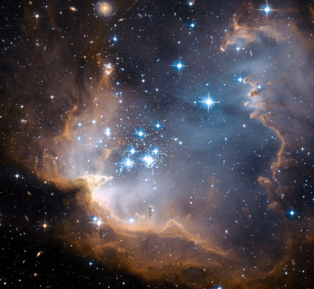 Bright blue stars are in a cluster at the center of the image. They are surrounded by a shell of brownish and burnt orange dust and gas. The shell has ridges that reach towards the stars like teeth. A number of more distant galaxies also appear in the background.