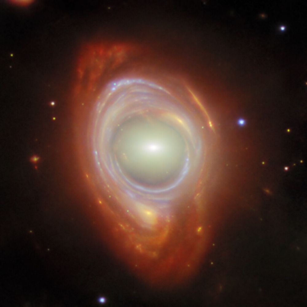 In the center is an elliptical galaxy, seen as an oval-shaped glow around a small bright core. Around this is wrapped a broad band of light, appearing like a spiral galaxy stretched and warped into a ring, with bright blue lines drawn through it where the spiral arms have been stretched into circles. A few distant objects are visible around the ring on a black background.