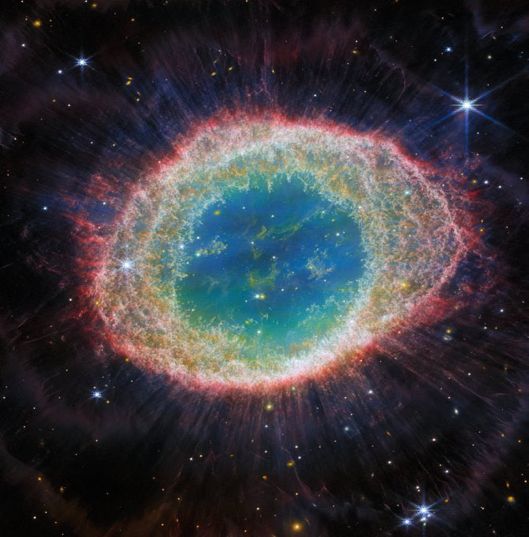 This image of the Ring Nebula, taken by Webb’s Near-Infrared Camera, appears as a distorted doughnut. The nebula’s inner cavity hosts shades of blue and green, while the detailed ring transitions through shades of orange in the inner regions and pink in the outer region. The ring’s inner region has distinct filament elements. Stars litter the scene, with a particularly prominent star with 8 long spikes in the top right corner.