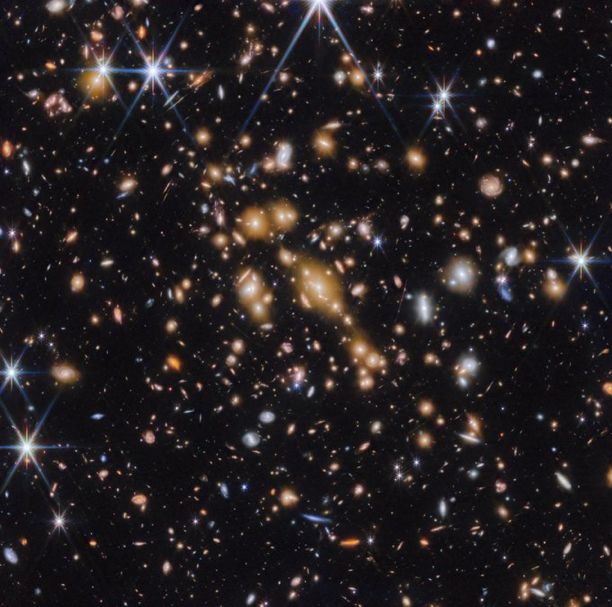 A field of galaxies on the black background of space. In the middle is a collection of dozens of yellowish galaxies that form a foreground galaxy cluster. Among them are distorted linear features, which mostly appear to follow invisible concentric circles curving around the center of the image. The linear features are created when the light of a background galaxy is bent and magnified through an effect called gravitational lensing. A variety of brightly colored, red and blue galaxies of various shapes are scattered across the image, making it feel densely populated.