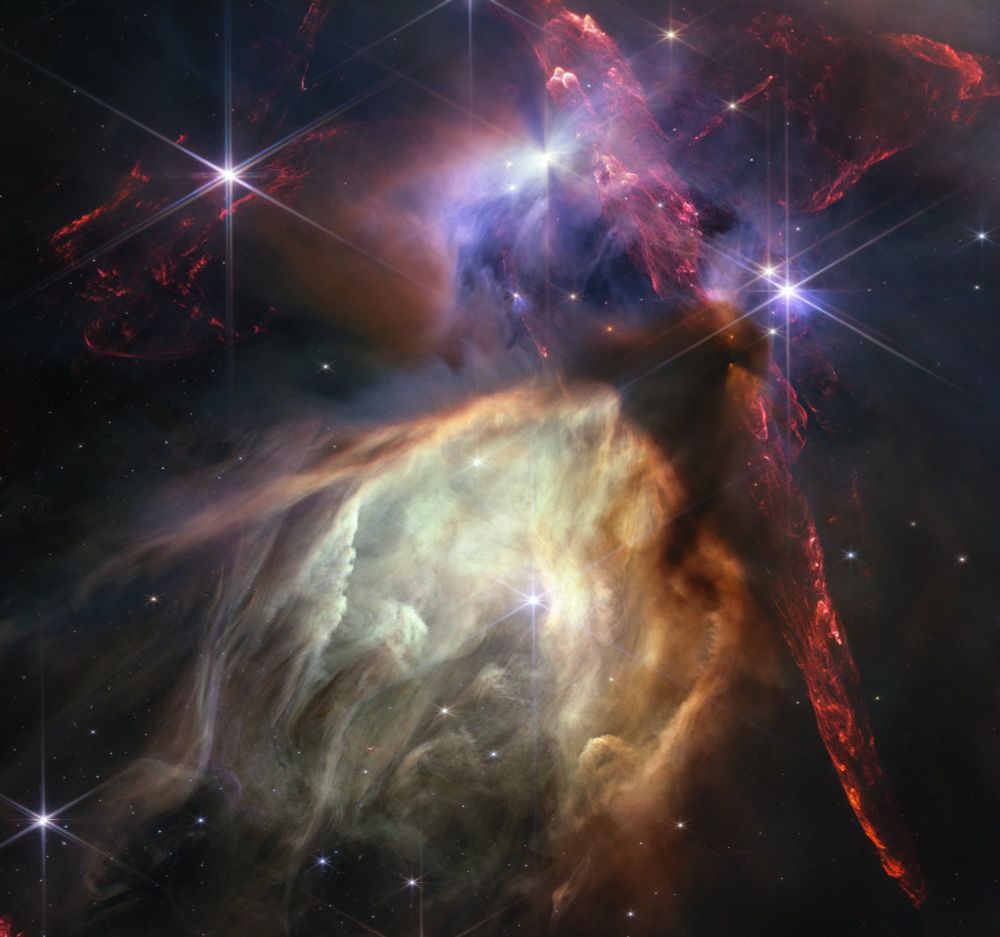 Red dual opposing jets coming from young stars fill the darker top half of the image. At bottom center is a glowing pale yellow, cave-like structure, its top tilted toward two o’clock, with a bright star at its center. The dust of the cave structure becomes wispy toward eight o’clock. Above the arched top of the dust cave, 3 groupings of stars with diffraction spikes are arranged. A dark cloud sits at the top of the arch of the glowing dust cave, with one streamer curling down the right-hand side. The dark shadow of the cloud appears pinched in the center, with light emerging in a triangle shape above and below the pinch, revealing the presence of a star inside the cloud. The largest jets of red material emanate from within this dark cloud, thick and displaying structure like the rough face of a cliff, glowing brighter at the edges. At top center, a star displays another, larger pinched dark shadow, this time vertically. To the left of this star is a more wispy, indistinct region.