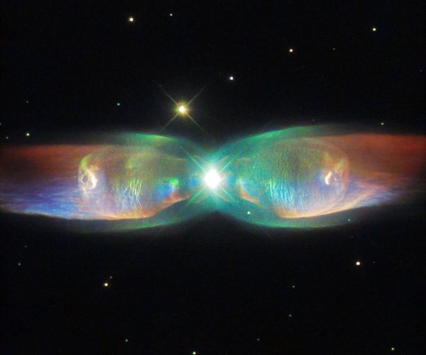 A bright white point of light is surrounded by glowing lobes of gas to the left and right, shining in green that extends into orange and blue. Distant stars dot the black background of space.