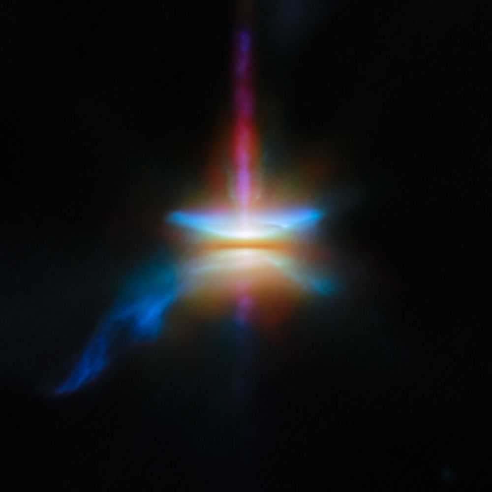 A close-in image of a protoplanetary disc around a newly formed star. Many different wavelengths of light are combined and represented by separate and various colors. A dark line across the center is the disc, made of opaque dust: the star is hidden in here and creates a strong glow in the center. A band going straight up is a jet, while other outflows form flares above and below the disc, and a tail coming off to one side.