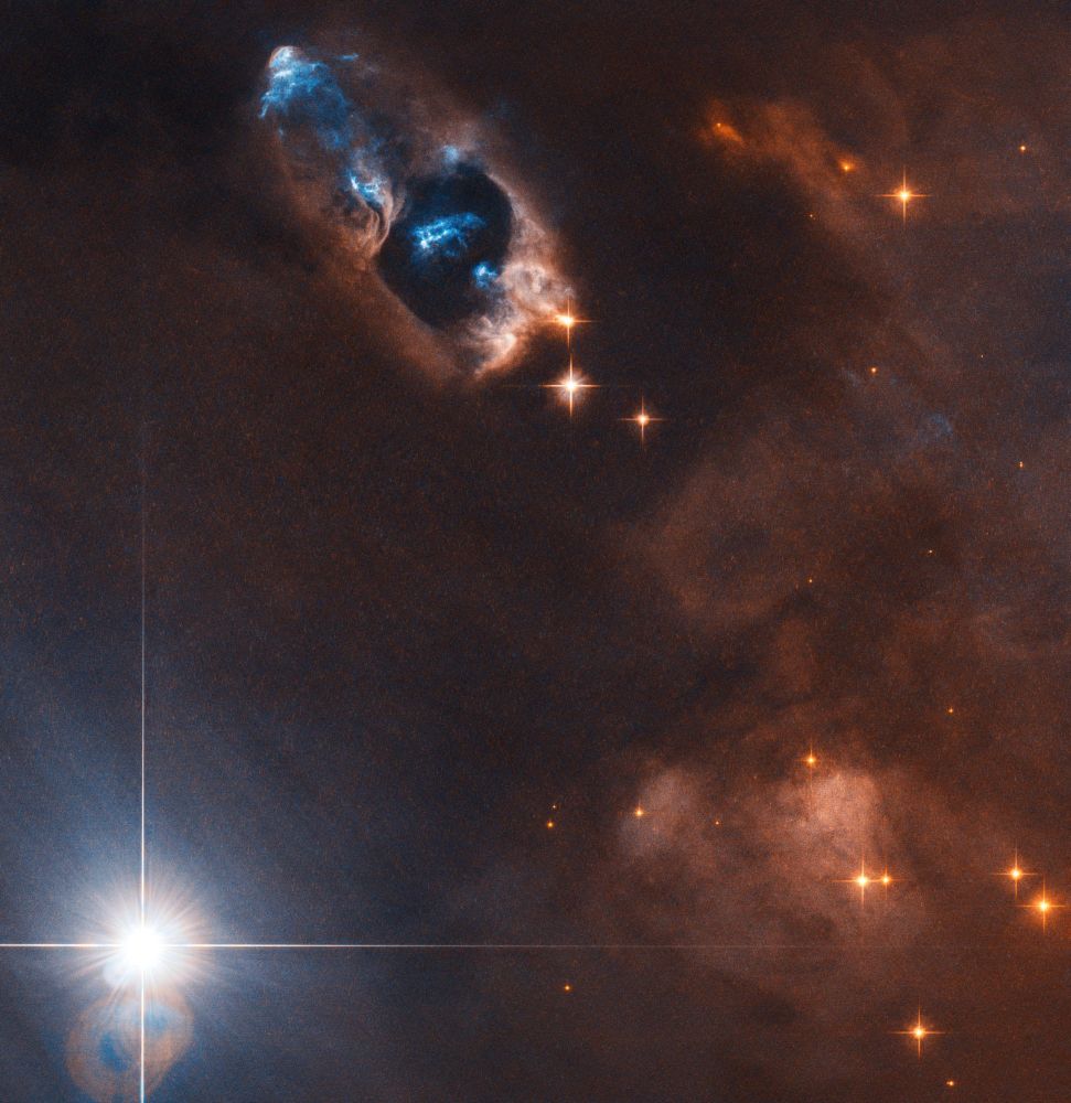 A ruddy nebula is pierced through with orange stars on the right side of the image, and a large white star with diffraction spikes shines at lower left. A cavity in the nebula is seen near upper left with bright blue wisps of gas throughout.