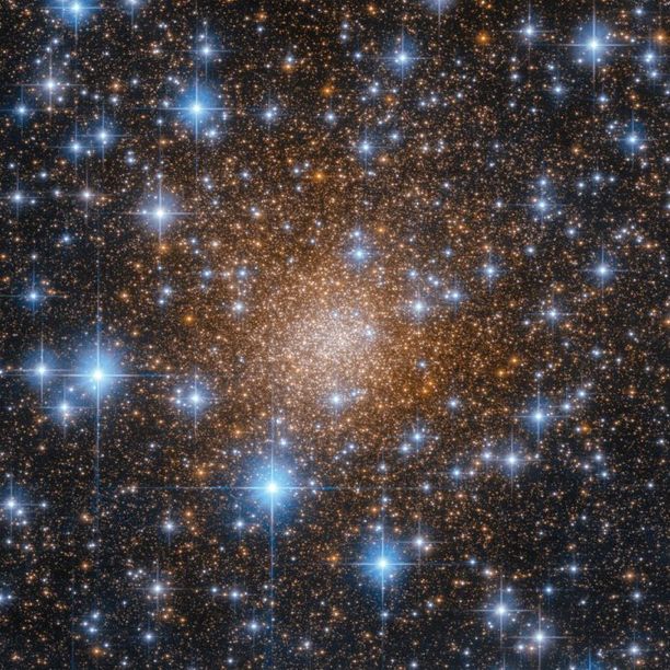 This star cluster features several orange stars packed together near the center and growing more sporadic toward the image's edges. In the foreground, bigger blue stars with diffraction spikes shine, all against a black backdrop of space.