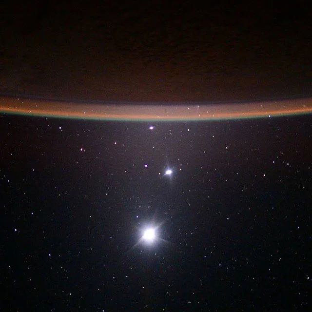 The starry background of space. At the top of the image is the orange glow of Earth’s atmosphere, with a few bright white dots forming a line above it. Those are the moon, Venus, and Jupiter.