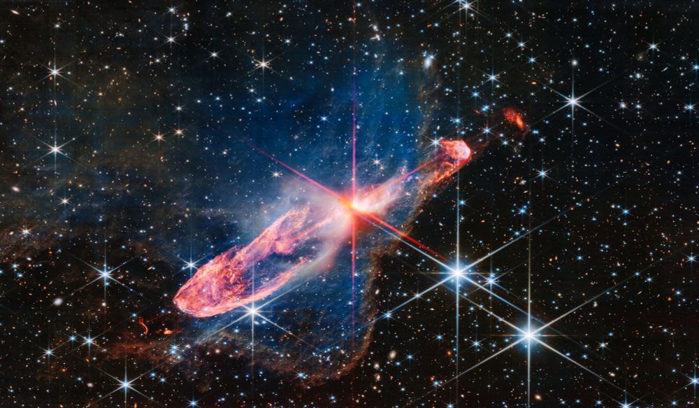 At the center of the image is a horizontal orange cloud known as Herbig-Haro 46/47 that is uneven with rounded ends, and tilted from bottom left to top right. In the middle of this cloud is a yellow-white blob with 8 reddish pink diffraction spikes piercing through it. The left lobe of the cloud is thicker. Just off the edge is a tiny red arc that curves in the opposite direction. The right lobe is thinner, and ends in a smaller orange semi-circle that has a faint purple outline. Just off the edge of this lobe is a slightly smaller orange sponge-like blob. A delicate, semi-transparent blue cloud known as a nebula drifts toward the top of the image and peters out toward the left of the frame. Toward the right and bottom, the nebula ends in a soft ridge set off in a translucent orange. The background is filled with stars and galaxies. Two foreground stars with blue diffraction spikes, seen in the bottom right corner, are especially prominent.