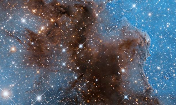 This Hubble view of a portion of the Carina Nebula has a bright, pale blue background dotted with shining white stars. Some foreground stars have diffraction spikes. A brown region of gas and dust snakes through the center of the image.
