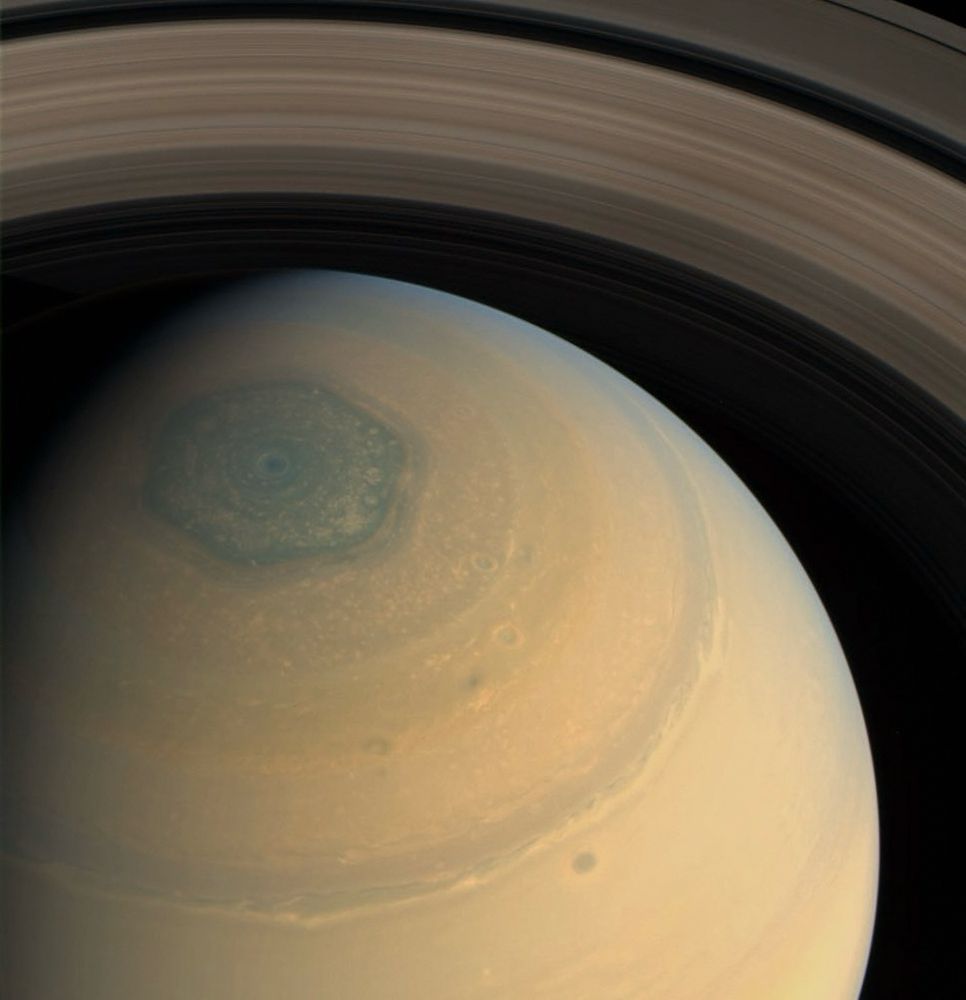 A close up of Saturn. Its banded atmospheric structure is visible in hues of gold, with its dark hexagon-shaped storm at its North Pole in a darker shade of blue and its iconic rings visible in the back. 