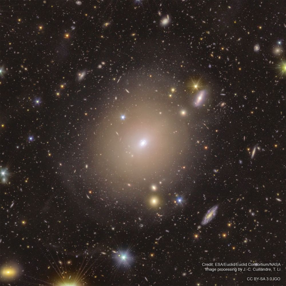 The large, hazy oval of elliptical galaxy NGC 6505 illuminates the center of this image from the European Space Agency's Euclid mission. At the heart of the galaxy is a small bright spot surrounded by a thin white circle, which is a magnified view of a much more distant galaxy. The view is also filled with countless colorful stars and galaxies of various sizes and shapes amidst the blackness of space. Thin spikes of light extend from the brightest stars due to the observatory’s optics. The image is watermarked “Credit: ESA/Euclid/Euclid Consortium/NASA, image processing by J.-C. Cuillandre, T. Li” and “CC BY-SA 3.0 IGO.”