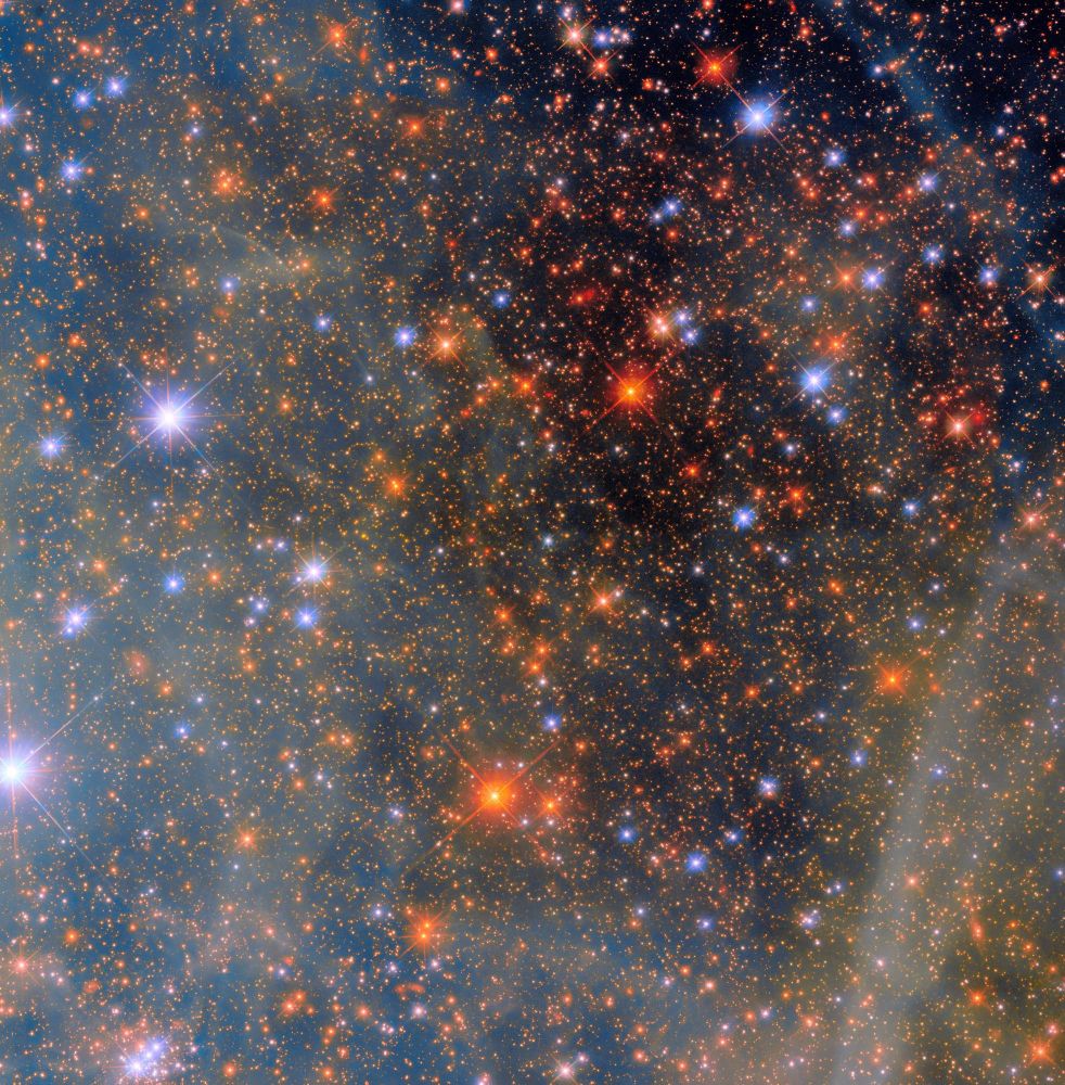 An area of space filled with stars. Most of the stars are small, distant dots in a range of orange colors; closer stars shine with a bright glow and hold four thin diffraction spikes around them. These closer stars appear both bluish and reddish. Clouds from a nebula cover the left half of the scene, giving it a blue-greenish cast. More clouds also drift over the black background of space on the right side of the image.