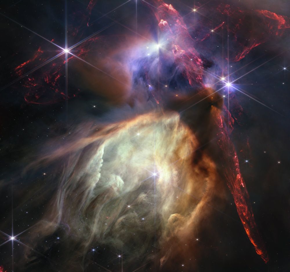 Red dual opposing jets coming from young stars fill the darker top half of the image. At bottom center is a glowing pale yellow, cave-like structure, its top tilted toward two o’clock, with a bright star at its center. The dust of the cave structure becomes wispy toward eight o’clock. Above the arched top of the dust cave, 3 groupings of stars with diffraction spikes are arranged. A dark cloud sits at the top of the arch of the glowing dust cave, with one streamer curling down the right-hand side. The dark shadow of the cloud appears pinched in the center, with light emerging in a triangle shape above and below the pinch, revealing the presence of a star inside the cloud. The largest jets of red material emanate from within this dark cloud, thick and displaying structure like the rough face of a cliff, glowing brighter at the edges. At top center, a star displays another, larger pinched dark shadow, this time vertically. To the left of this star is a more wispy, indistinct region.