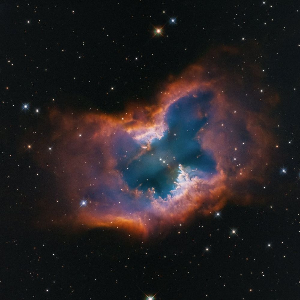 A glowing nebula shines against the black background of space. The planetary nebula is shaped like a single macaroni noodle, with its edges pointed up, but its edge-on central torus is semi-transparent in the middle. The top and bottom edges are thick and orange. The center is semi-transparent blue and green. The wider central region looks roughly like a moth, also filled with semi-transparent blue and green. There are two pinpoint-like white stars with diffraction spikes toward the center. Immediately below them, slightly toward the right, is a smaller blue orb, a central star. The next layer of gas and dust is whiter, with some thicker pillars that look like they are rising up at bottom center. The color fades into reds and purples, and then to orange. The orange outline fades toward the left and right, but appears thicker at top and bottom. The background is speckled with pinpoints of light, most of which look like dots. Larger orbs appear with four bright spikes.