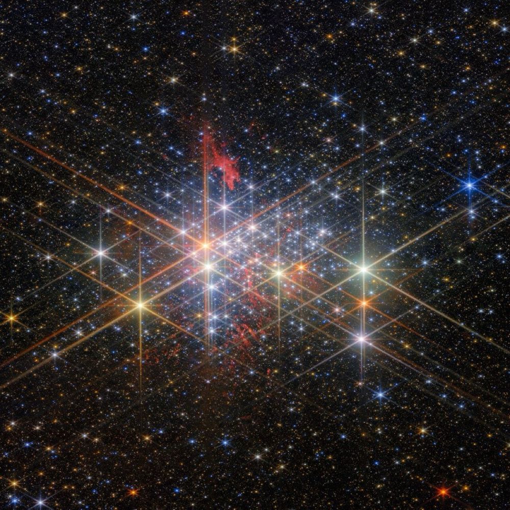 A wider field view of Westerlund 1. A dense cluster of bright stars, each with six large and two small diffraction spikes, due to the telescope’s optics. They have a variety of sizes depending on their brightness and distance from us in the cluster, and different colours reflecting different types of star. Patches of billowing red gas can be seen in and around the cluster, lit up by the stars. Small stars in the cluster blend into a background of distant stars and galaxies on black.