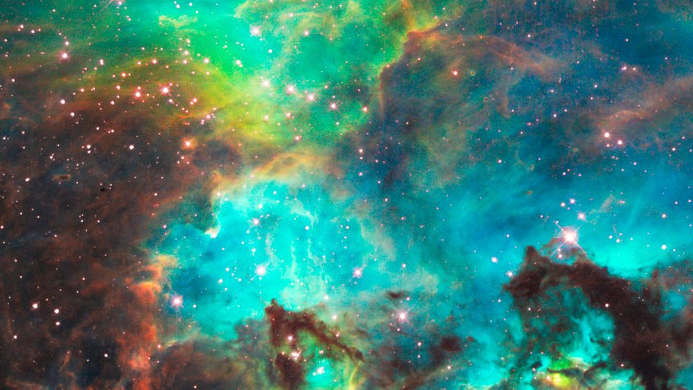 Intricate clouds of gas and dust fill the image. Dark brown dust is concentrated on the left side of the image, along with the lower portions of the rest of the view. Glowing green and blue gas fills the rest of the image, with pink stars visible throughout.
