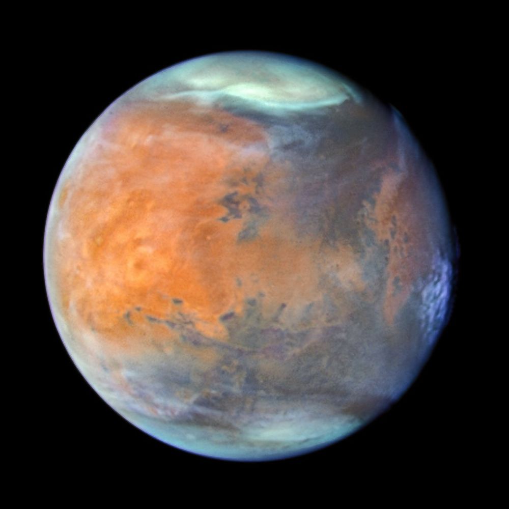 The Red Planet appears brightest orange in the left half. Darker surface features are noticeable on the lower half of the planet. These have a mix of orange, blue, and gray hues. At the top and bottom, white regions mark the planet’s polar caps. The entire limb of the planet, its visible edge, has a blue hue. The blue doesn’t form an even circle at the edges, and appears thinner toward the left and right, and thicker in some areas.