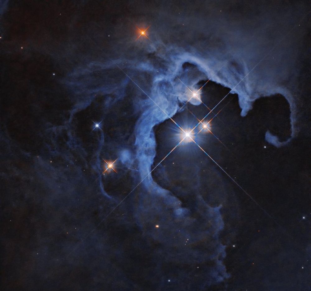 Three bright stars with diffraction spikes shine near the center-right of the image, illuminating nearby clouds that glow in pale blue. The clouds darken at the edges of the image, and are dotted with smaller stars, some also with diffraction spikes.

image credit: NASA, ESA, G. Duchene (Universite de Grenoble I); image processing: Gladys Kober (NASA/Catholic University of America)