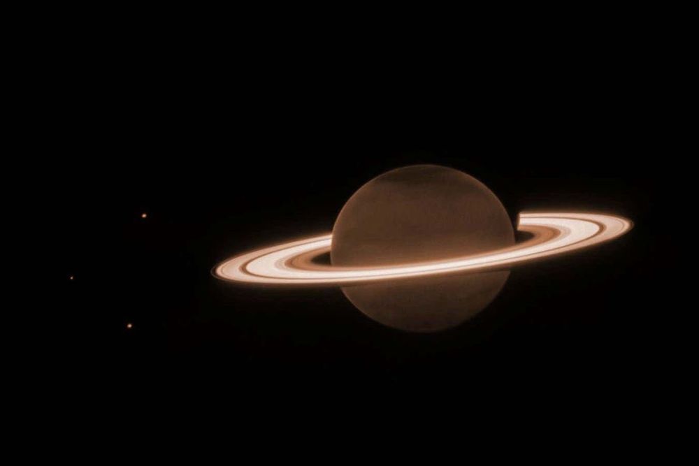 The background is mostly dark. At the center is a dark orange-brown circle, surrounded by several blazing bright, thick, horizontal whiteish rings. This is Saturn and its rings. There are three tiny dots in the image—one to the upper left of the planet, one to the direct left of the planet, and the lower left of the planet. These are three of Saturn’s moons: Dione, Enceladus, and Tethys, respectively. There is a slightly darker tint at the northern and southern poles of the planet. The rings surrounding Saturn are mostly broad, with a few singular narrow gaps between the broader rings. There is an innermost, thicker ring, and next to that is a brighter, wider ring. Traveling farther outward, there is a small dark gap before another thicker ring. In the thicker ring, there is a narrow faint band. There is then an outermost, faintest, thinnest ring.