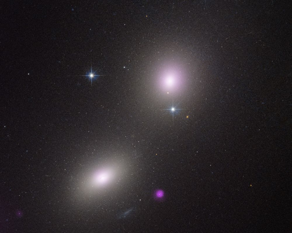 Two elliptical galaxies at upper right and lower left appear as fuzzy white ovals. At bottom center is a fuzzy purple blob. The main image also has two bright stars with prominent diffraction spikes. The background of space is black and is speckled with tiny white dots.