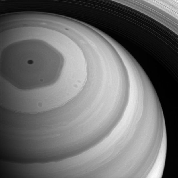 A close up of Saturn in black and white. Its banded atmospheric structure is visible with its dark hexagon-shaped storm at its North Pole. 