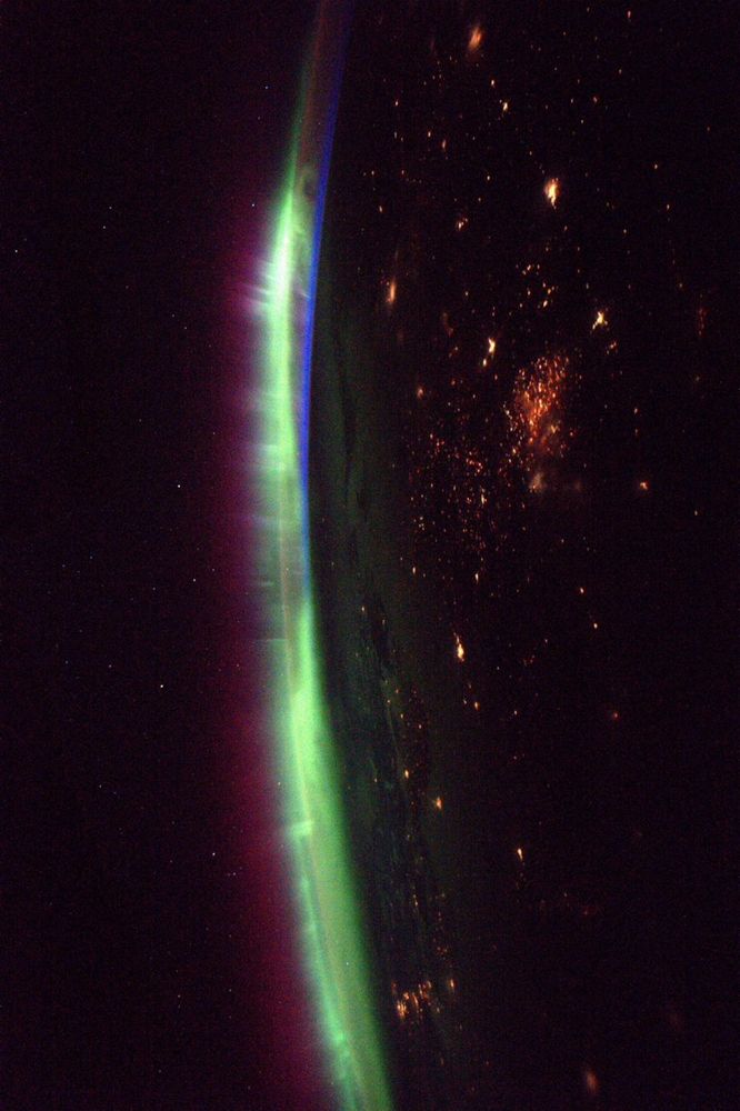 A picture of the green and red aurora above earth from space. It is nighttime on Earth, some lights are visible. Some stars are visible in the background.