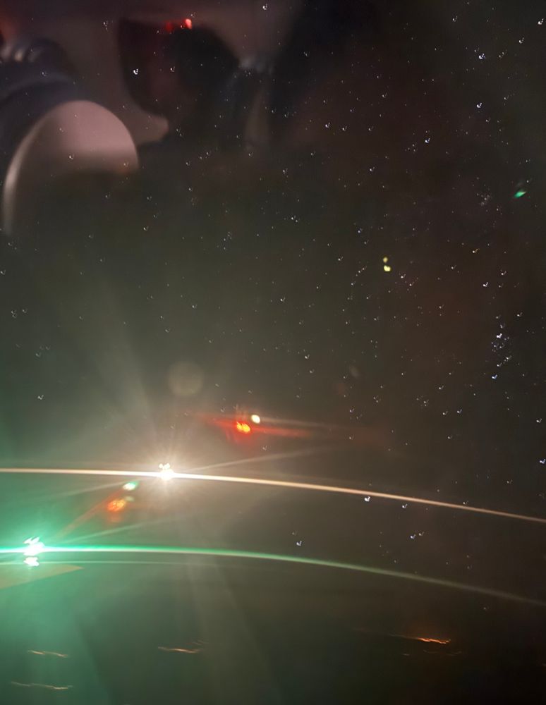 A long exposure photo of the night sky from my flight. There are many stars in the sky and green and white streaks from the plane lights. There is also a small reflection in the upper left from the plane.