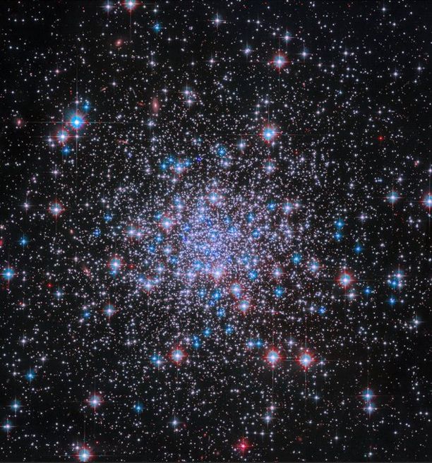 Thousands of bright stars shine against black space, more tightly condensed near the image’s center. The stars glint in shades of white, blue, and red, and diffraction spikes are visible around the foreground stars.