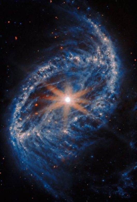 A spiral galaxy, seen close-up. Its core is a round spot that glows intensely bright, crowned by eight long and spikes that extend across the galaxy, artefacts of the telescope’s structure. Its disc is an oval shape with edges made of very thick and cloudy arms of gas and dust, mostly blue but paler and brighter around patches of stars. Wisps of darker dust also fill the inner disc and swirl off the ends of the arms.