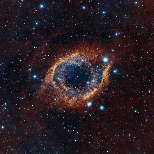The Helix Nebula appears as an eye in the center of the image. The outer layers appears yellow, while the inner layers are a darker blue color. There are many blue stars visible around the nebula. The background of the image shows dark red space.