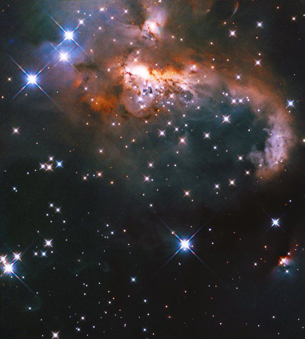 Three bright, blue-white stars at upper left mark the beginning of a bright and dusty reddish-brown, orange, and white swathe of glowing gas and dust, with its upper portions loosely resembling a snowman with a scarf and top hat. The rest of the image is dark and full of sparkling stars, many with diffraction spikes.