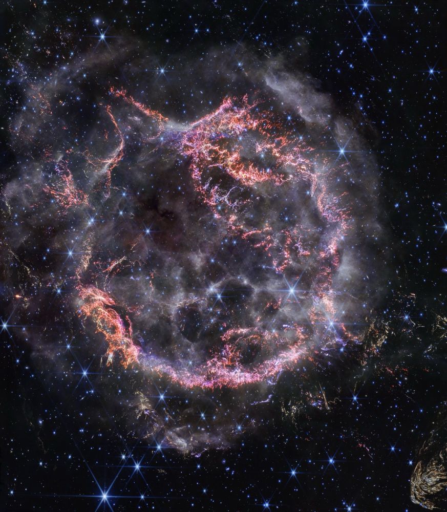 Cassiopeia A, a circular cloud of gas and dust with complex structure. The inner shell is made of bright pink and orange filaments studded with clumps and knots. Around the exterior of the inner shell, particularly at the upper right, there are curtains of wispy gas that look like campfire smoke. The white smoke-like material also appears to fill the cavity of the inner shell, featuring structures shaped like large bubbles. Around and within the nebula, there are various stars seen as points of blue and white light. Outside the nebula, there are also clumps of yellow dust, with a particularly large clump at the bottom right corner that appears to have very detailed striations.