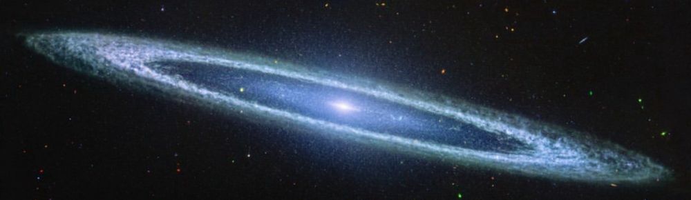 The galaxu is a very oblong, blue disk that extends from left to right at an angle (from about 10 o’clock to 5 o’clock). The galaxy has a small bright core at the centre. There is a clear inner disk that has speckles of stars scattered throughout. The outer disk of the galaxy is whiteish-blue, and clumpy, like clouds in the sky.