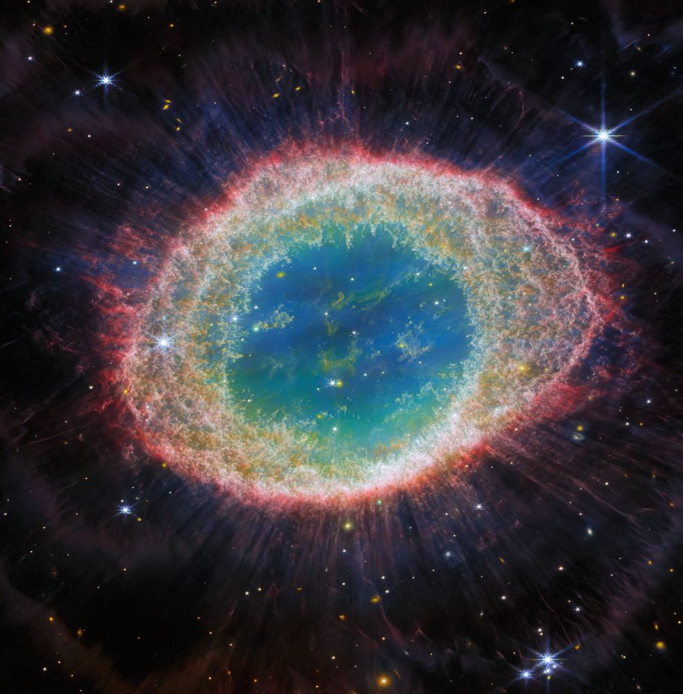 This image of the Ring Nebula, taken by Webb’s Near-Infrared Camera, appears as a distorted doughnut. The nebula’s inner cavity hosts shades of blue and green, while the detailed ring transitions through shades of orange in the inner regions and pink in the outer region. The ring’s inner region has distinct filament elements. Stars litter the scene, with a particularly prominent star with 8 long spikes in the top right corner.