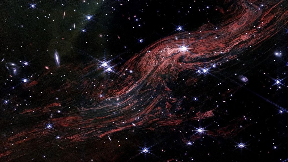 An infrared light echo traveling from the lower left through the upper right of the image. The image is speckled with dozens of white stars, some showing Webb’s signature 8-point diffraction spikes, against the black background of space. The image also shows tightly packed, glowing red filaments that resemble muscle fibers or wood grain.⁣