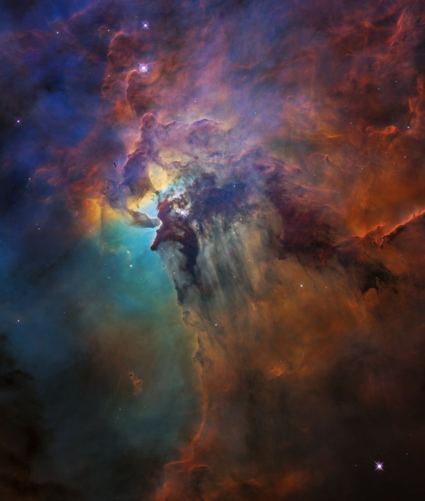Colorful clouds of gas and dust crowd the image. Deep red and orange clouds surround the image's edges, with some violet in the upper left portion. Near the left middle, a region of teal and yellow shines.