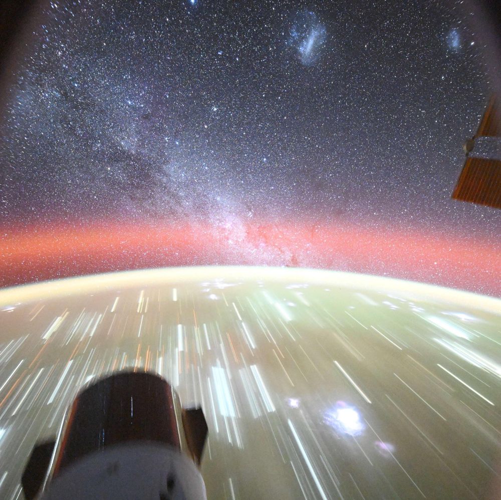 A long exposure image of the Earth from orbit showing the Dragon capsule, the city lights appearing like streaks with the green and orange atmospheric glow, many stars in the background of space with the streak of the Milky Way and both Magellanic Clouds as large circular blobs.