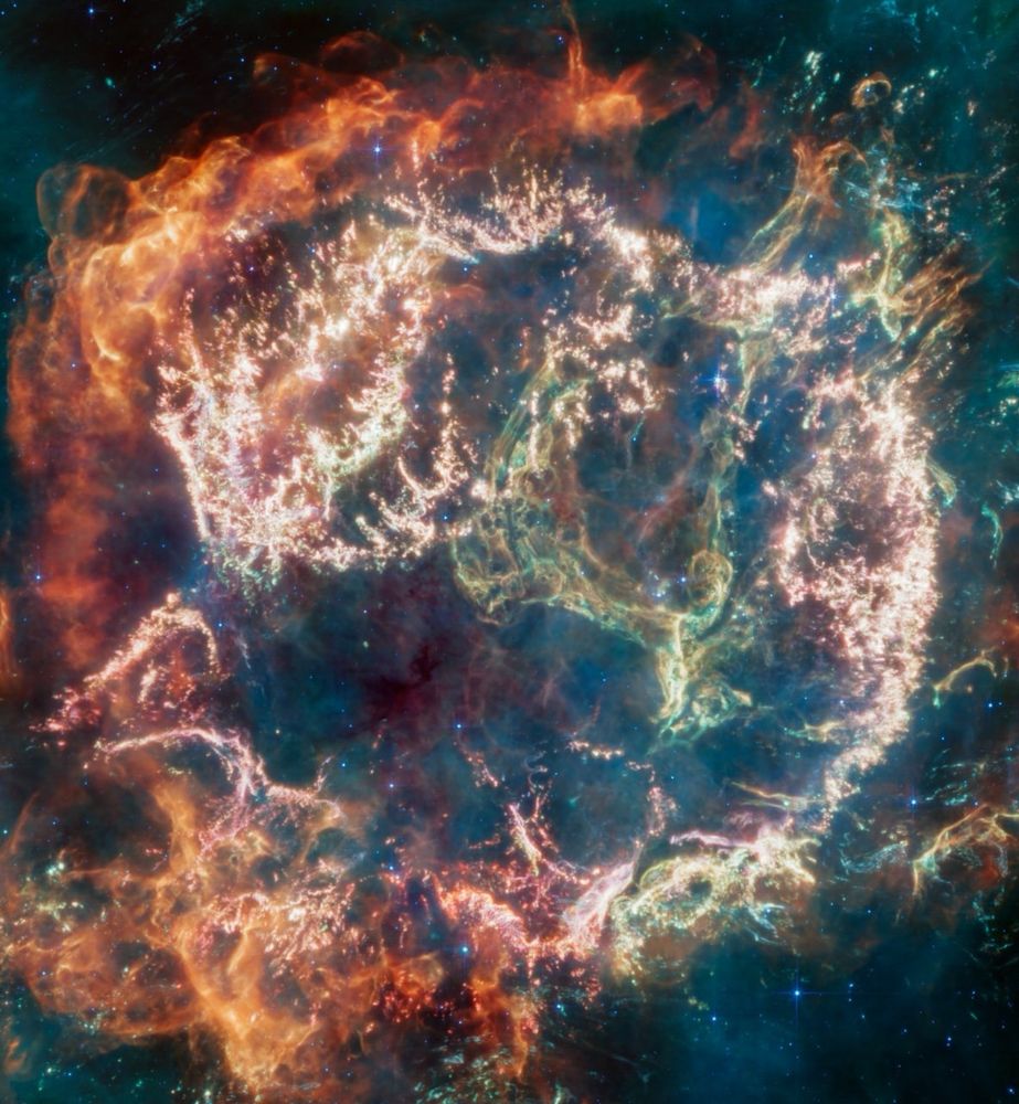 A circular-shaped nebula with a complex structure. On the circle’s exterior, particularly on the left side of the image, are curtains of material glowing orange like fire. Interior to this outer shell is a ring of mottled filaments in bright pink, studded with clumps and knots. A greenish loop extends from the upper right of the ring into the central cavity. Translucent wisps of blue, green, and red appear throughout the image.