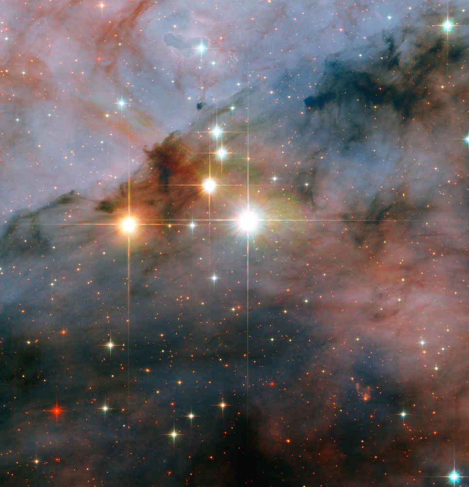 A small grouping of large stars shines brightly with diffraction spikes, against a background full of smaller stars, glowing reddish and blue gas, and dark dust.