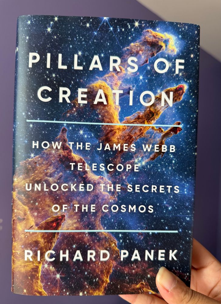 My hand holding up a book titled: “Pillars of Creation: How the James Webb Telescope Unlocked the Secrets of the Cosmos” by Richard Panek. The cover is the JWST image of the Pillars of Creation.