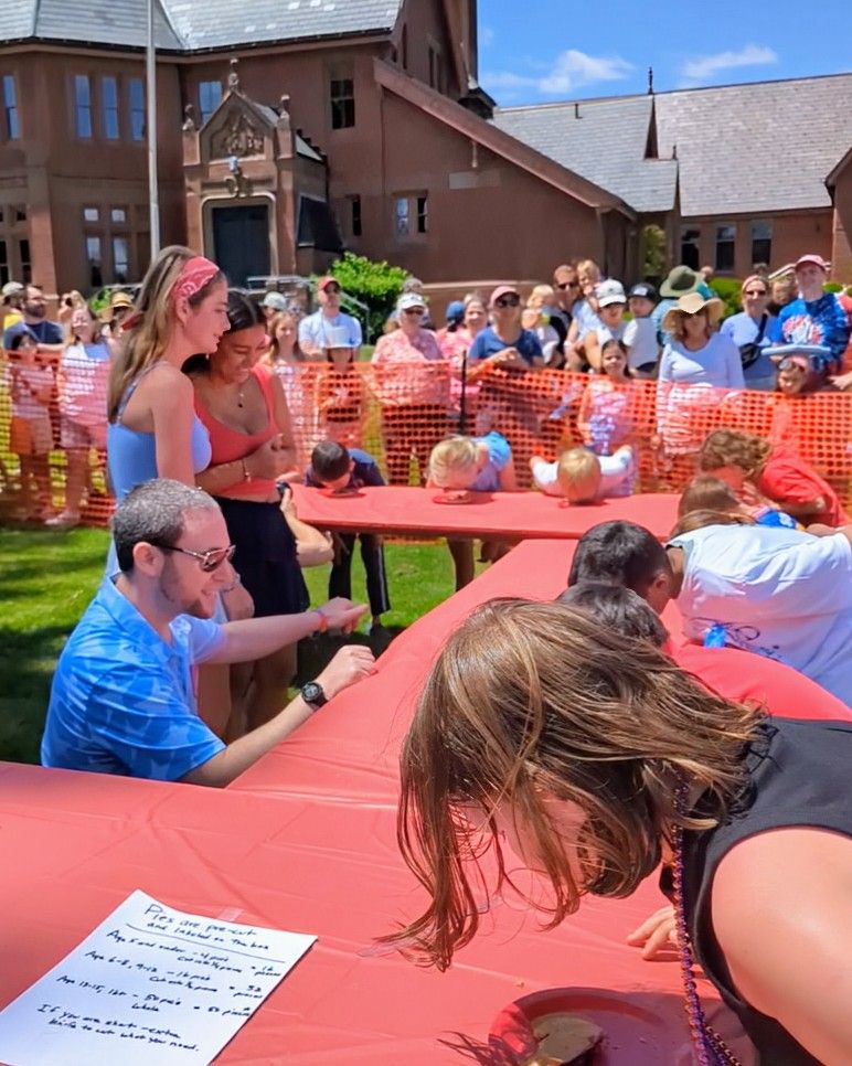 Alex kneeling to get a better view of the pies. Around the table are a bunch children, heads down, eating the pies.
