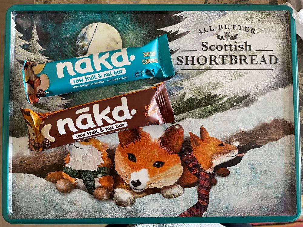 Picture of two nakd bars, one salted caramel and one cocoa orange, on top of an old shortbread snack tin with pictures of foxes on it 