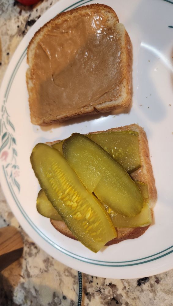 a pickle sandwich during assembly. two slices of bread, one has peanut butter and pickles and the other has just peanut butter.