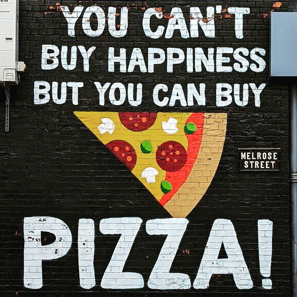 A wall painted back.  Written in bold, white capital letters is "YOU CAN'T BUY HAPPINESS BUT YOU CAN BUY PIZZA! 
There is a picture of a cartoon slice of pizza above the word pizza. The cartoon pizza shows a cheese and tomato base with pepperoni, mushrooms and possibly peppers.  There is a street sign on the wall also that says MELROSE STREET.