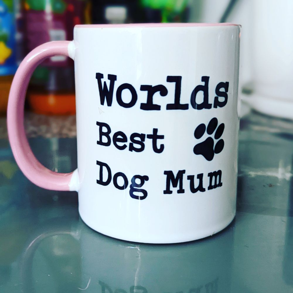 White mug with a pink handle sitting on a kitchen counter. Written on the mug in black text is "Worlds Best Dog Mum" with a pawprint.