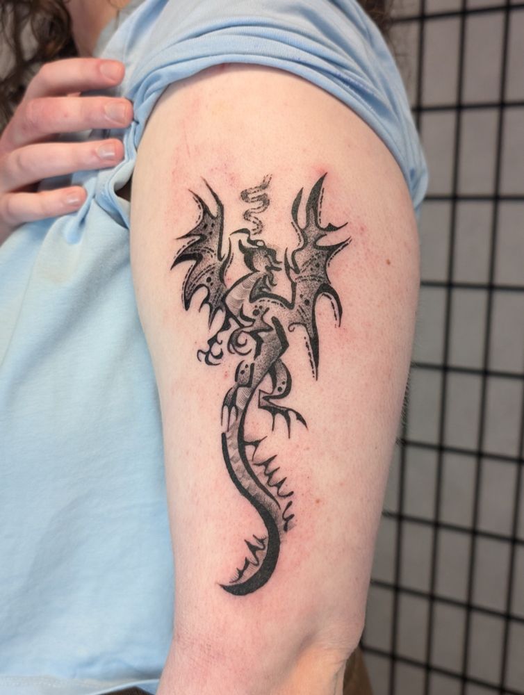 a black dragon tattoo on a woman's upper arm. the dragon is flying up the arm and there is a small trail of smoke coming out of its nose.