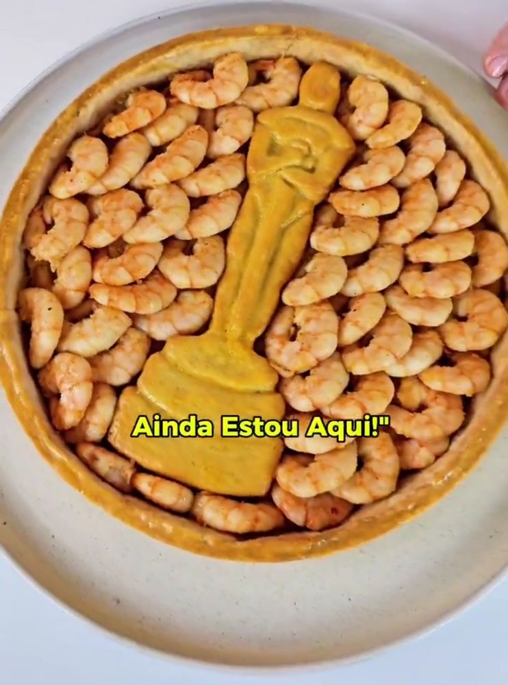 Massa de empadão já assada sobre um prato de cerâmica bege clarinho, dentro da massa foi colocado uma imagem da estatueta do Oscar feita com mais massa de empadão assado bem douradinha, ao redor camarões cozidos foram perfeitamente colocados, está escrito AINDA ESTOU AQUI na legenda da tela 