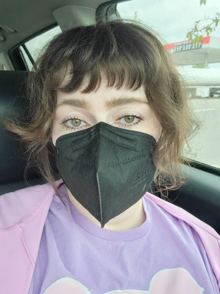 it me, in a kroger parking lot, in a car. gas station is visible in the left window (to the right in the pic). i'm wearing a black KN95 mask, a lilac gloomy bear shirt, and a bright pastel pink jacket.