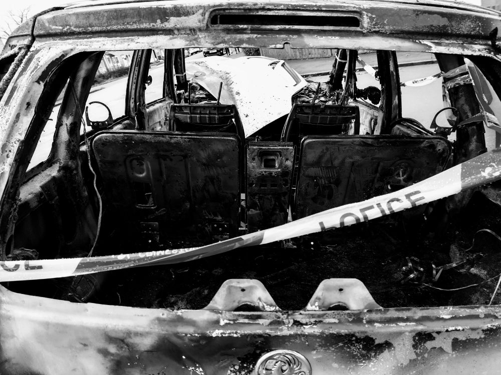 Black and white photo of a car, stolen, burned out and left behind. Photo is through the rear smashed window with some police tape across the frame.