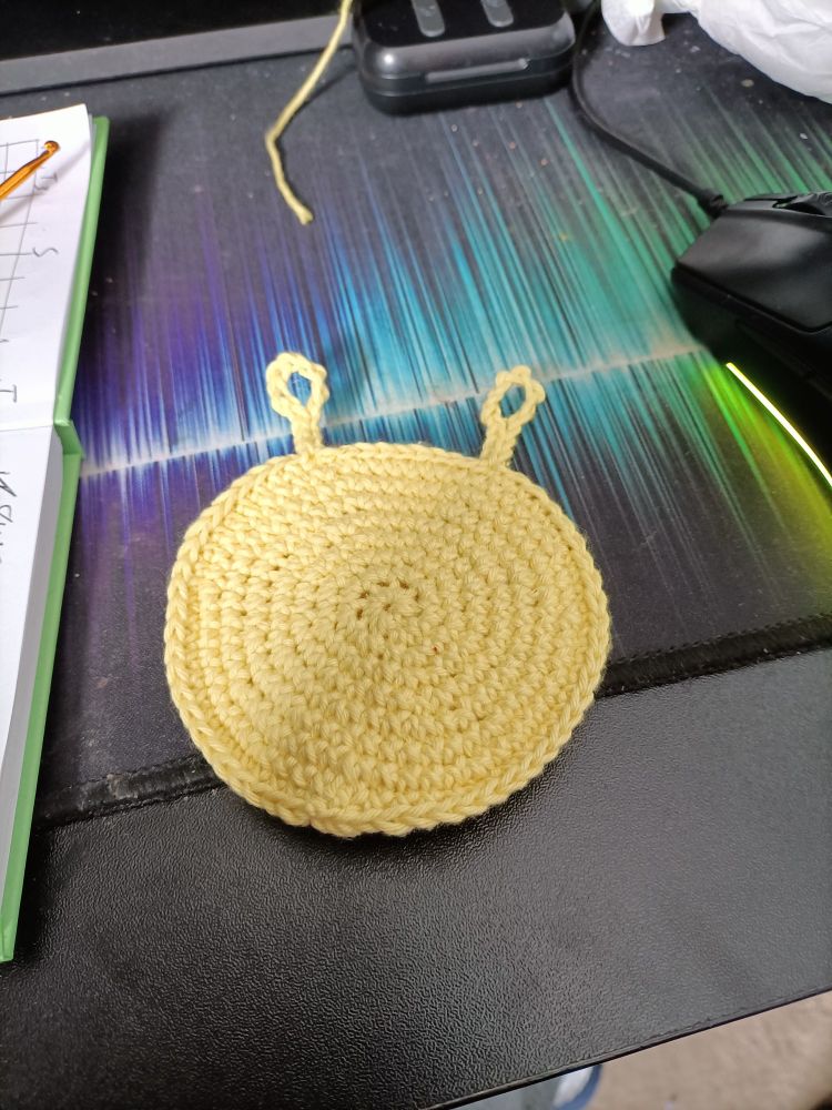 Same desk as image two, showing the yellow tummy of the in progress turtle. It is a yellow circle with two straps at the top ending in loops. A computer mouse, set of wireless earbuds and a notebook and crochet hook are visible in frame.